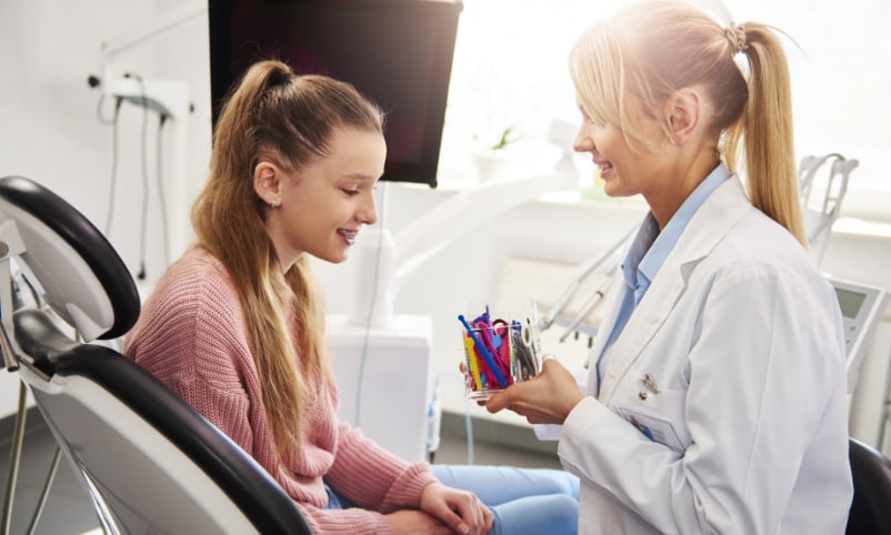 A female dentist is talking to a young girl in a dental chair.