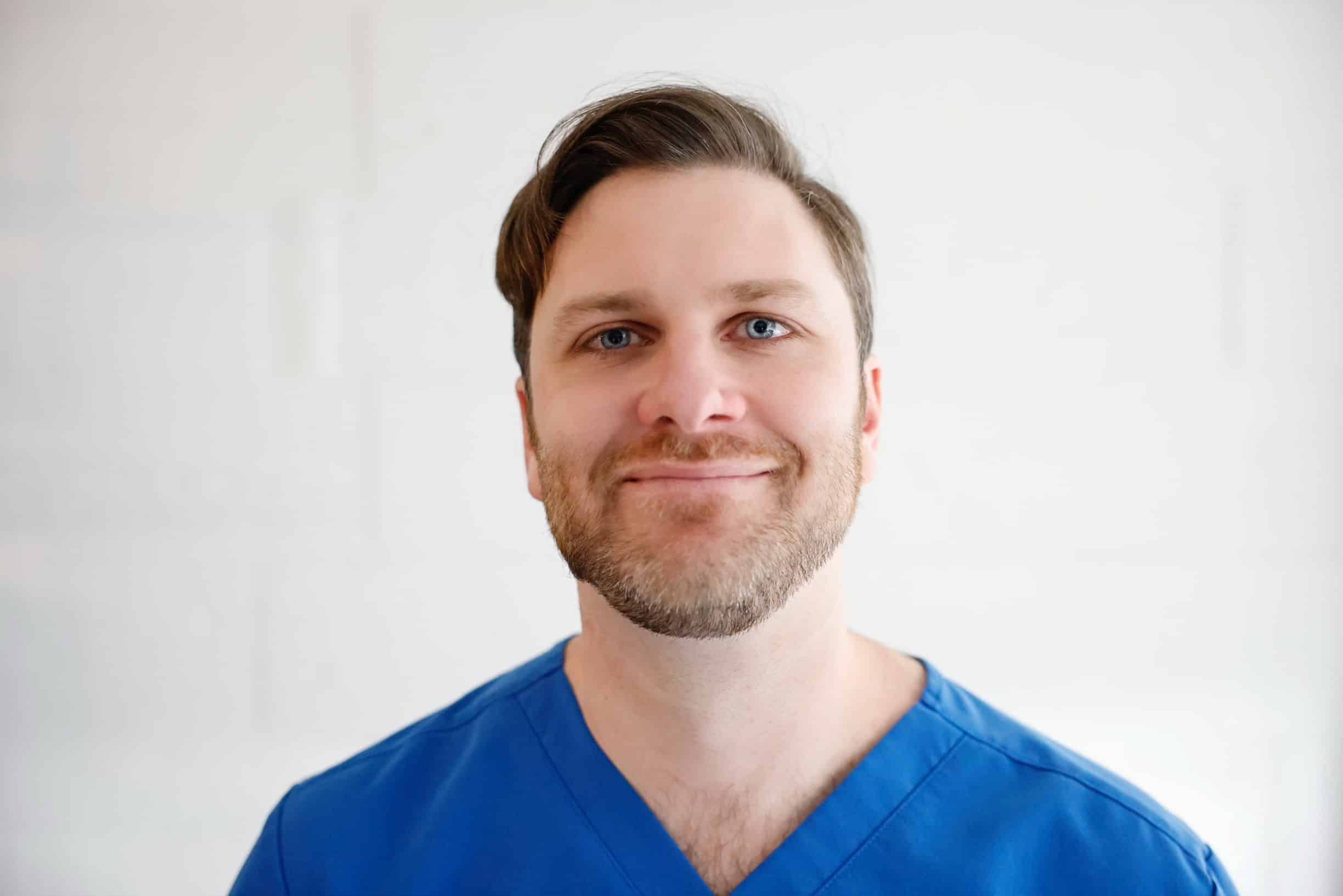A man in a blue scrub shirt smiles for the camera.